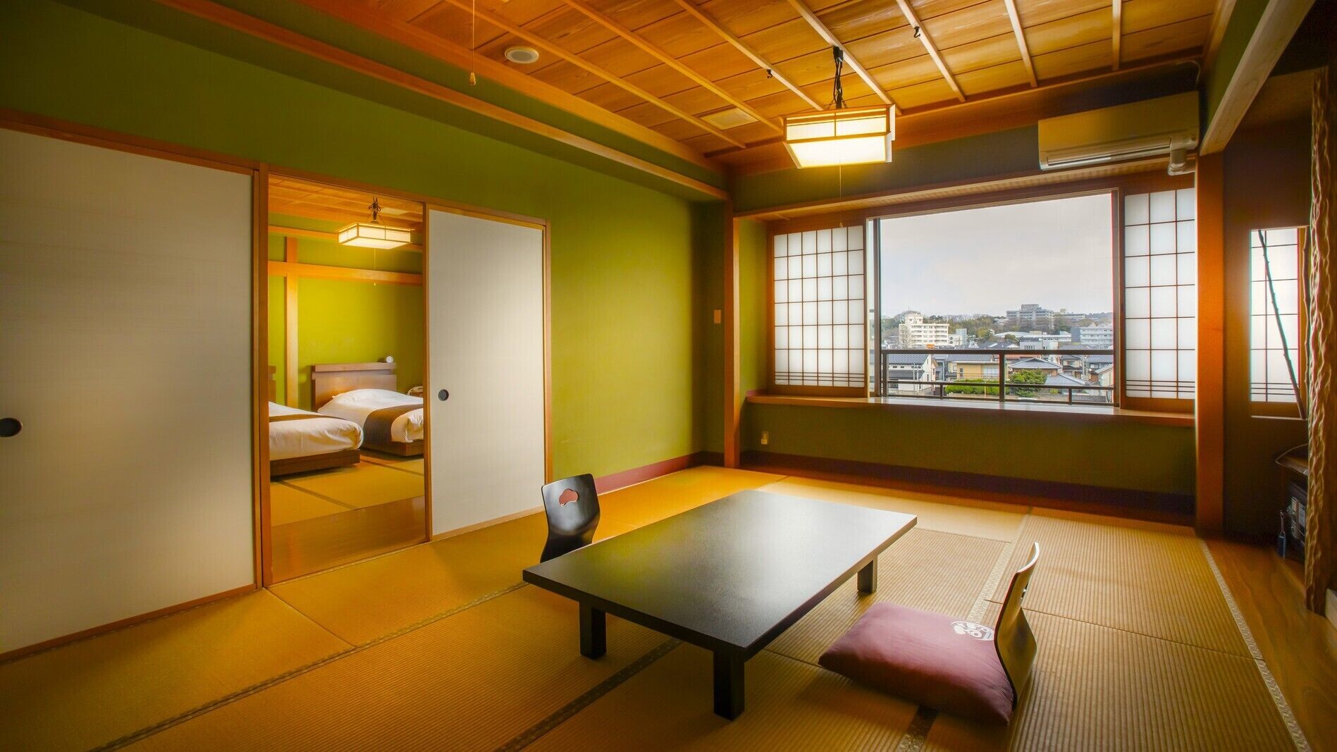 Ein traditioneller japanischer Raum mit Tatami-Boden, einem niedrigen Tisch, einer gepolsterten Sitzbank und einer Schiebetür zu einem Schlafzimmer mit einem Bett.