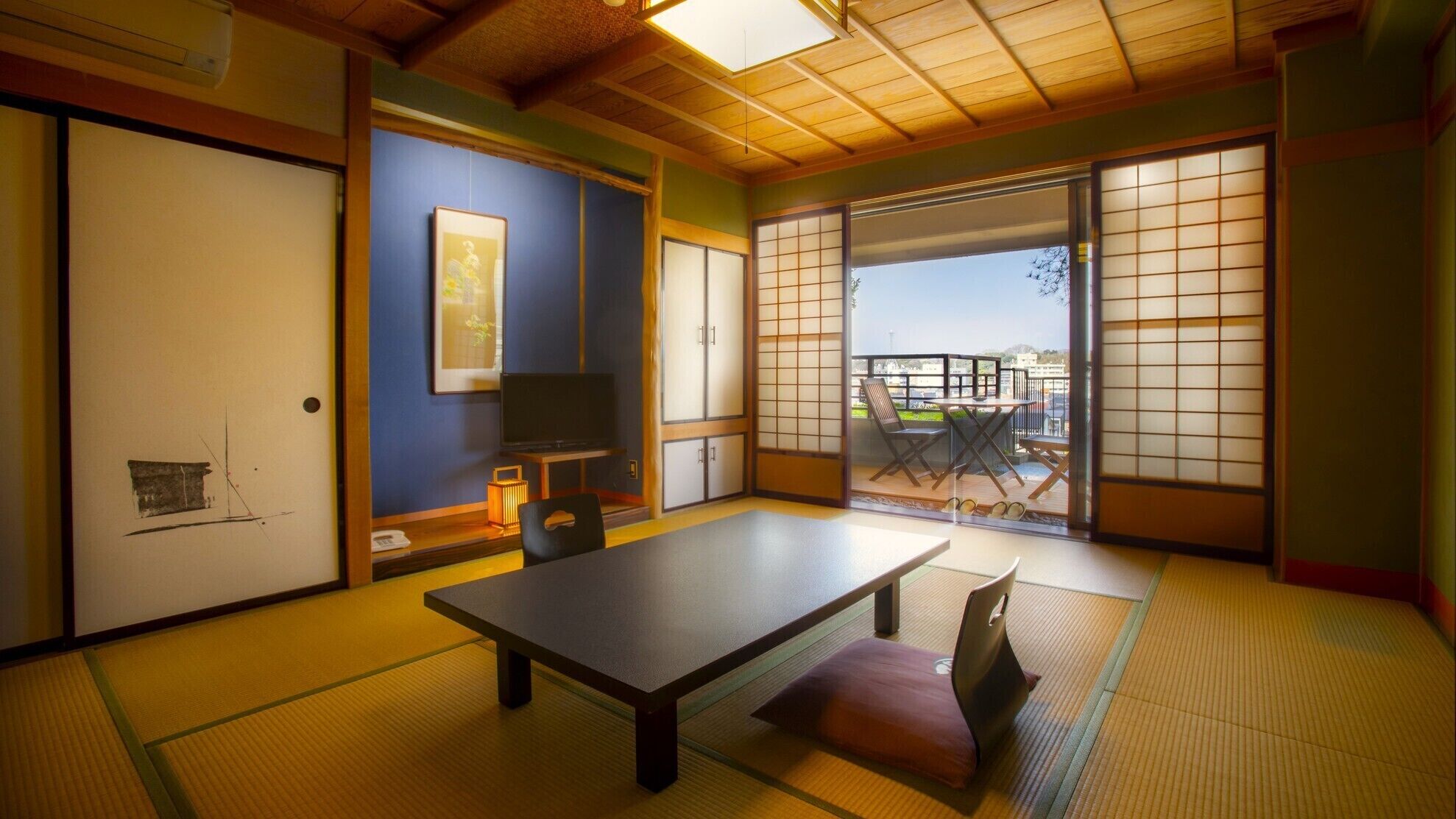 Ein traditioneller japanischer Raum mit Tatami-Bodenbelag, einem niedrigen Tisch, einem Fernseher und Schiebetüren, die zu einem Balkon im Freien führen.