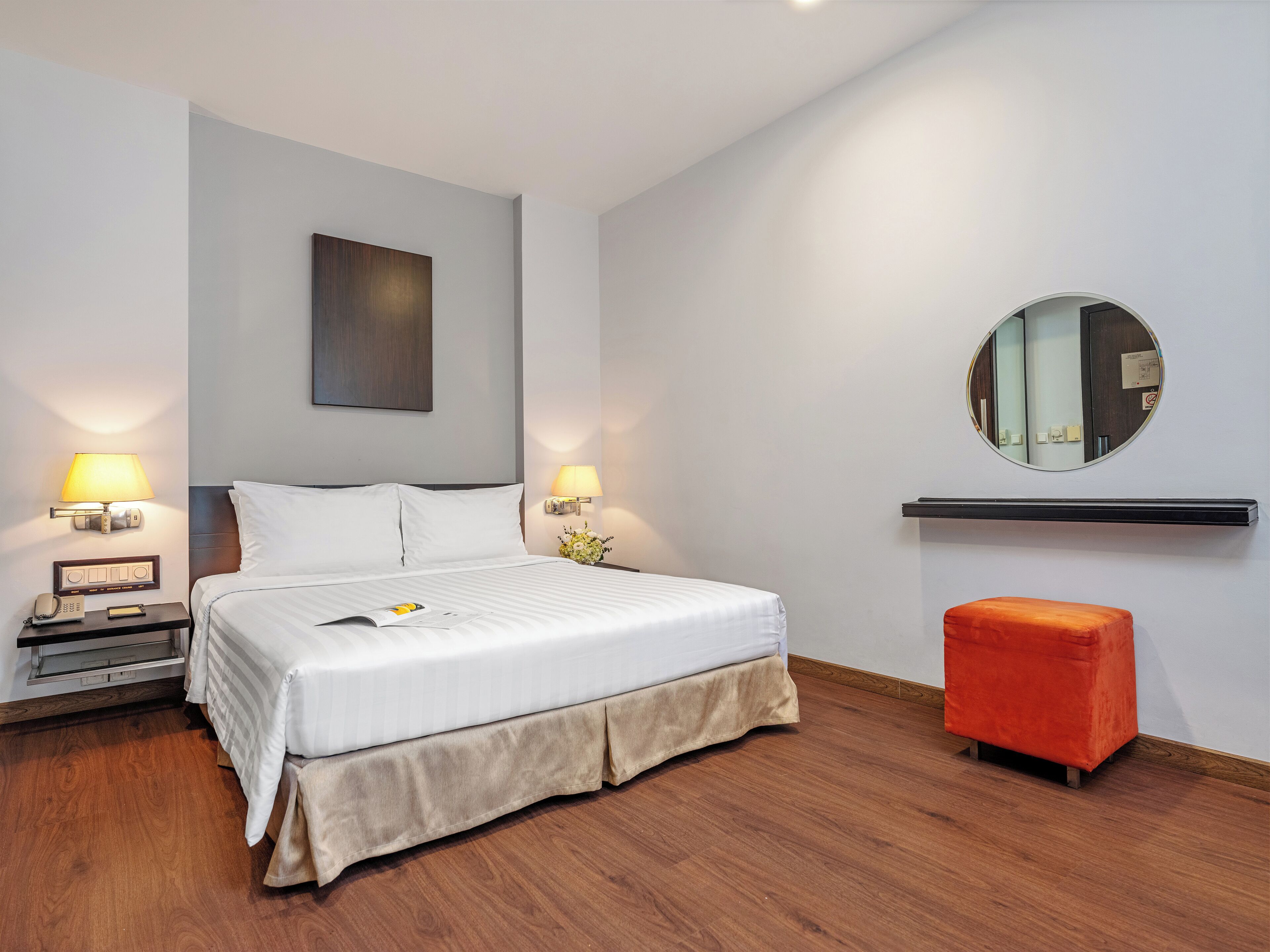 A hotel room with a bed, bedside lamps, a nightstand, a mirror, and a small orange ottoman.
