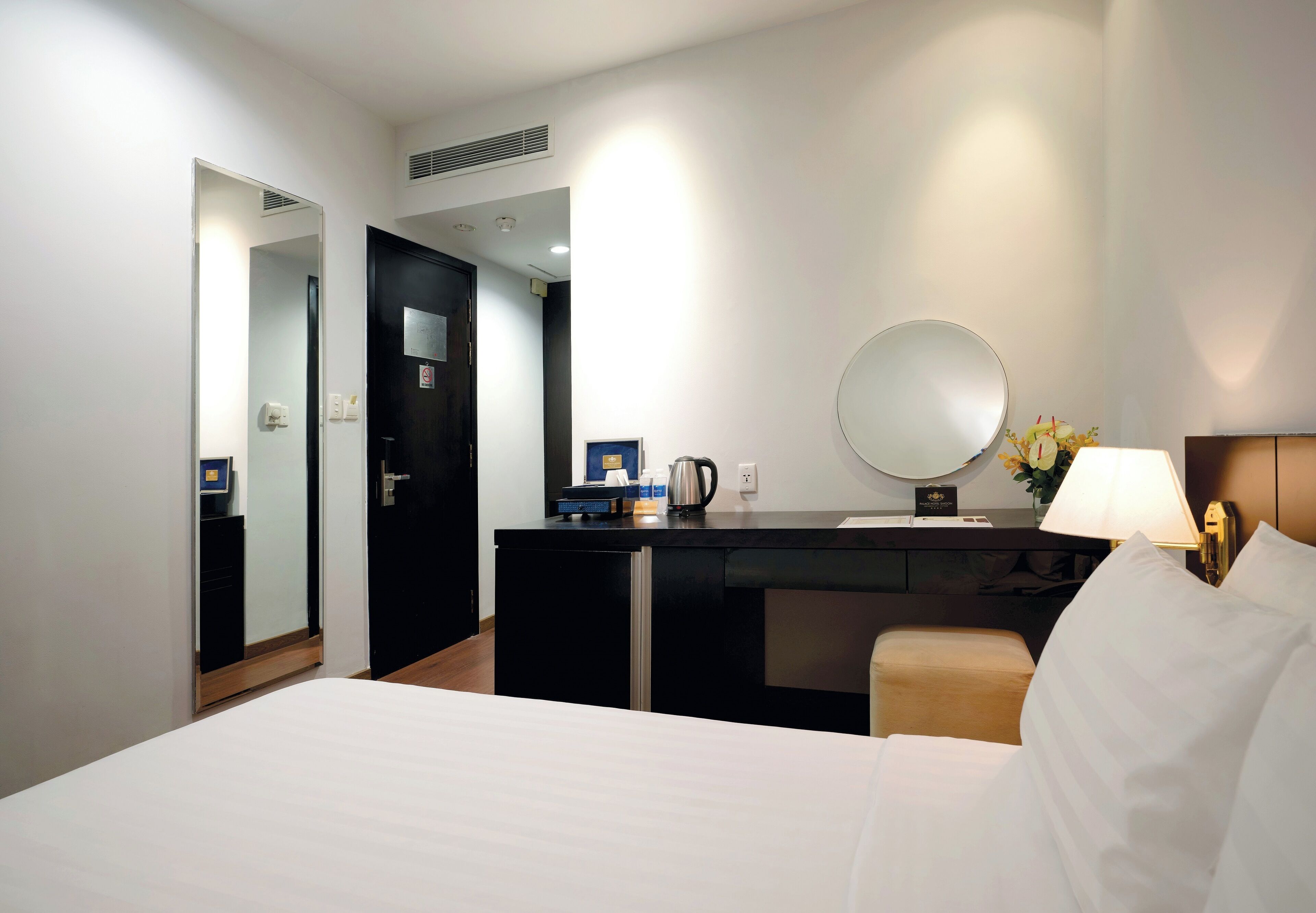 Une chambre d’hôtel avec un lit, un bureau doté d’un miroir, une lampe et une petite table sur laquelle se trouve une cafetière.