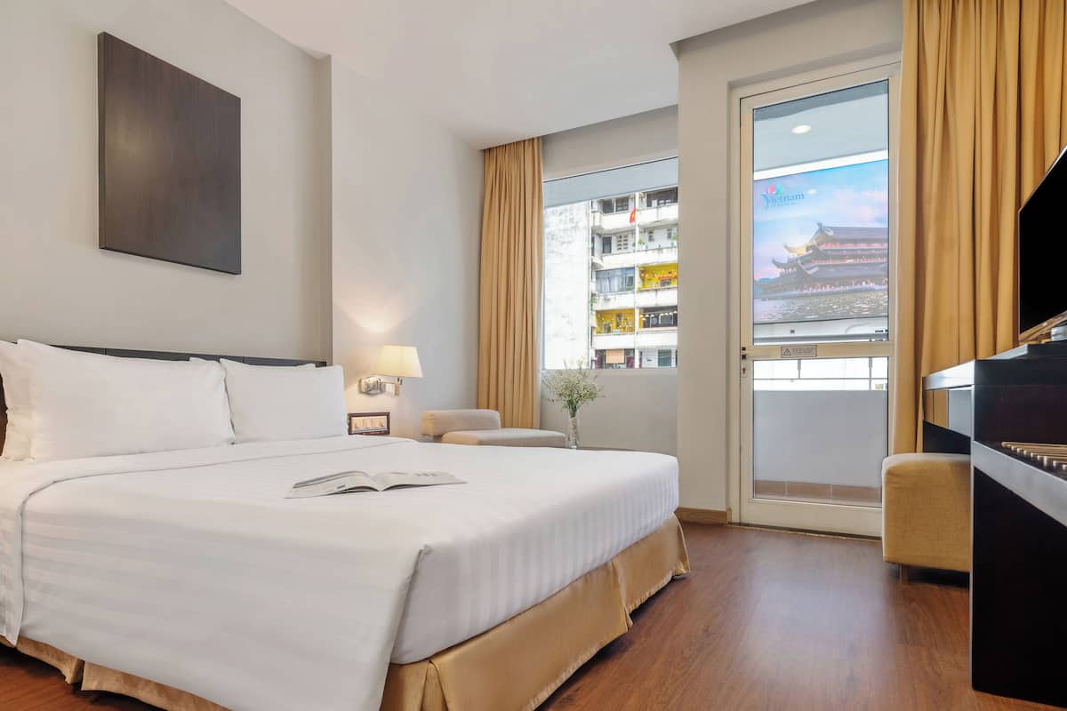 Ein Hotelzimmer mit einem großen Bett, einem Schreibtisch mit Klavier, einem Fenster mit Blick auf ein Gebäude und einer Wandlampe.