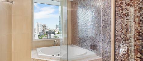 A modern bathroom with a glass-enclosed jacuzzi, a tiled shower area, and a view of the cityscape through a large window.