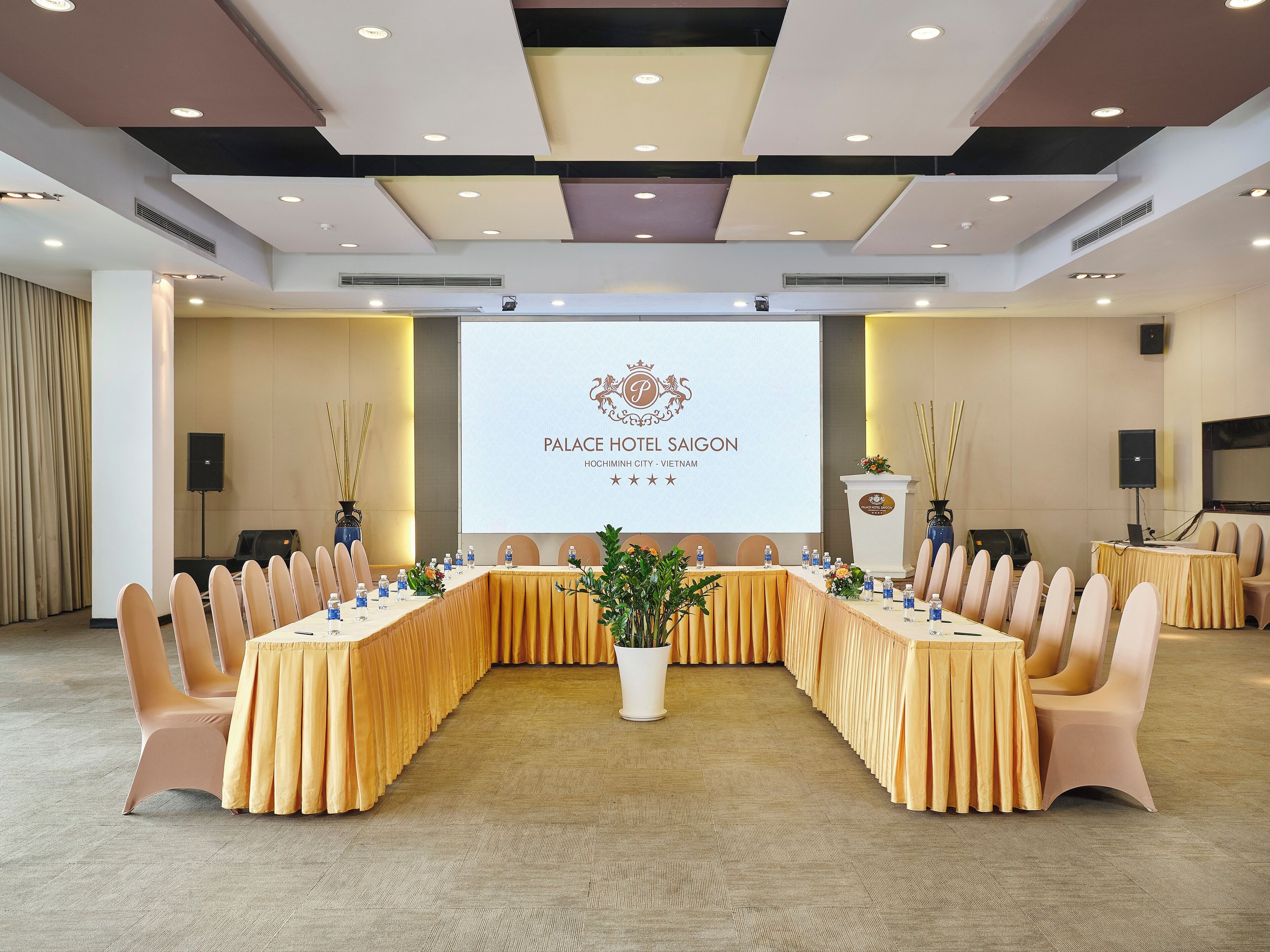 Un salón de conferencias con una larga mesa, sillas, un atril y una pantalla que muestra el nombre del hotel.