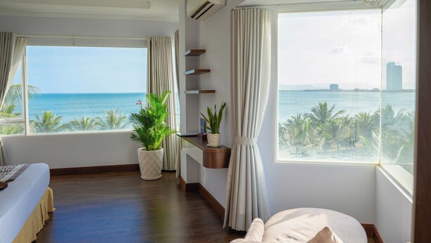 A hotel room with a large window offering a view of the sea and palm trees, a bed, a bedside table with a plant, and a small shelf unit. - Saigon Ninh Chu Hotel & Resort (Ninh Chu)
