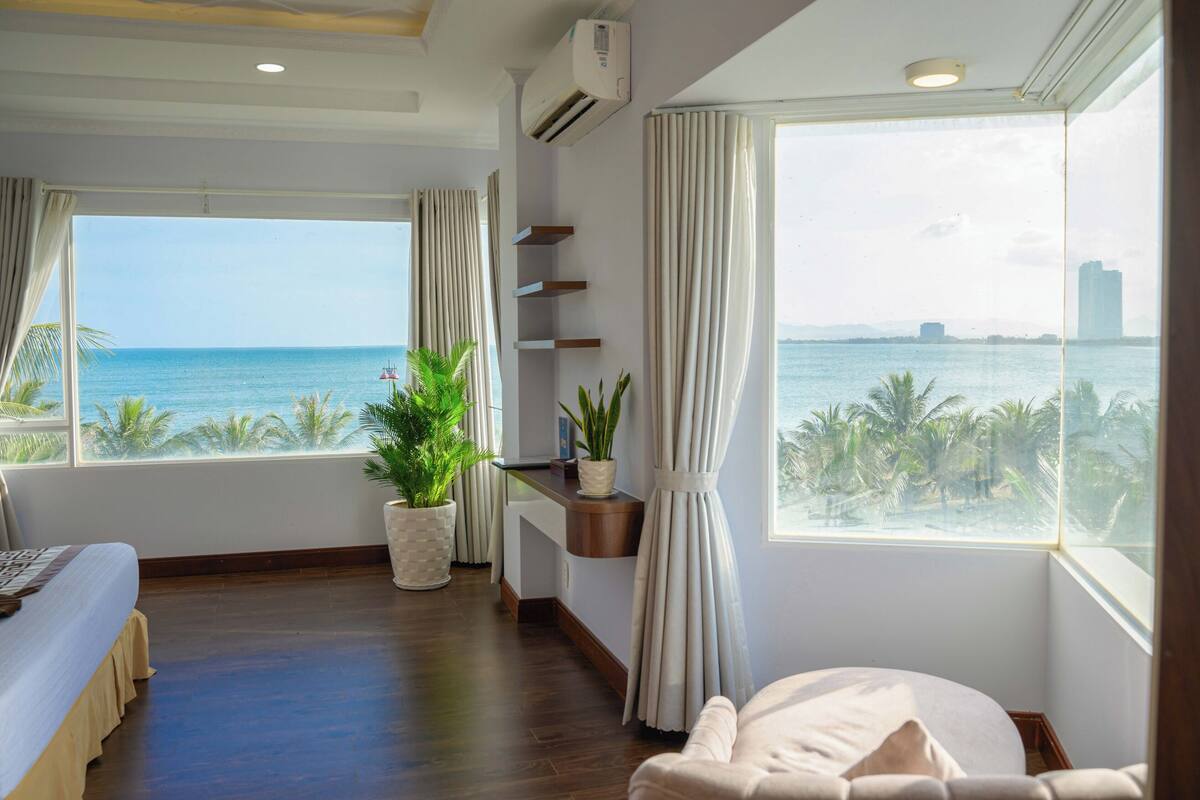 A hotel room with a large window offering a view of the sea and palm trees, a bed, a bedside table with a plant, and a small shelf unit.