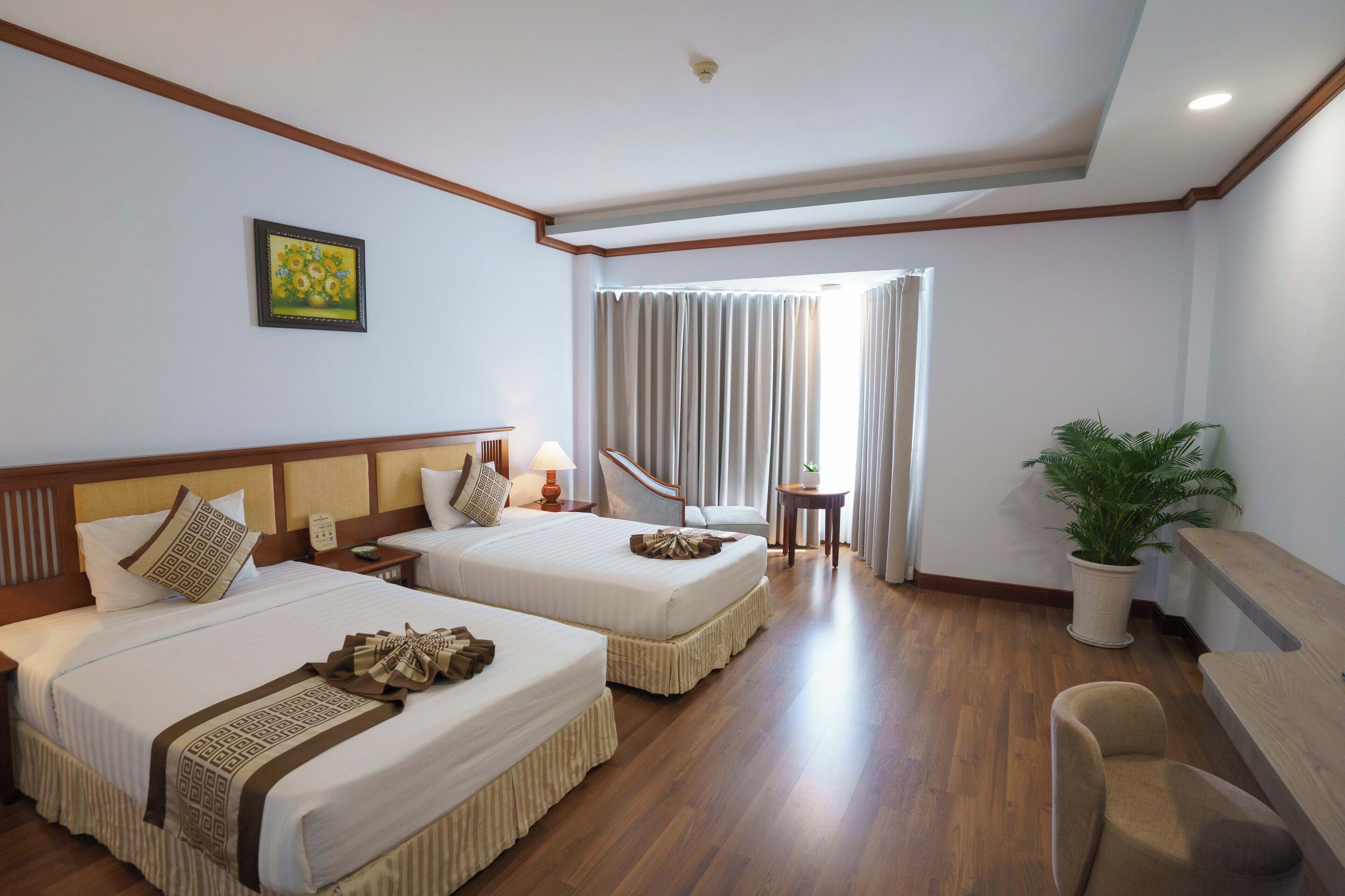 A hotel room with two beds, a desk, a chair, a potted plant, and a window with curtains.