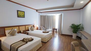 A hotel room with two beds, a desk, a chair, a potted plant, and a window with curtains.