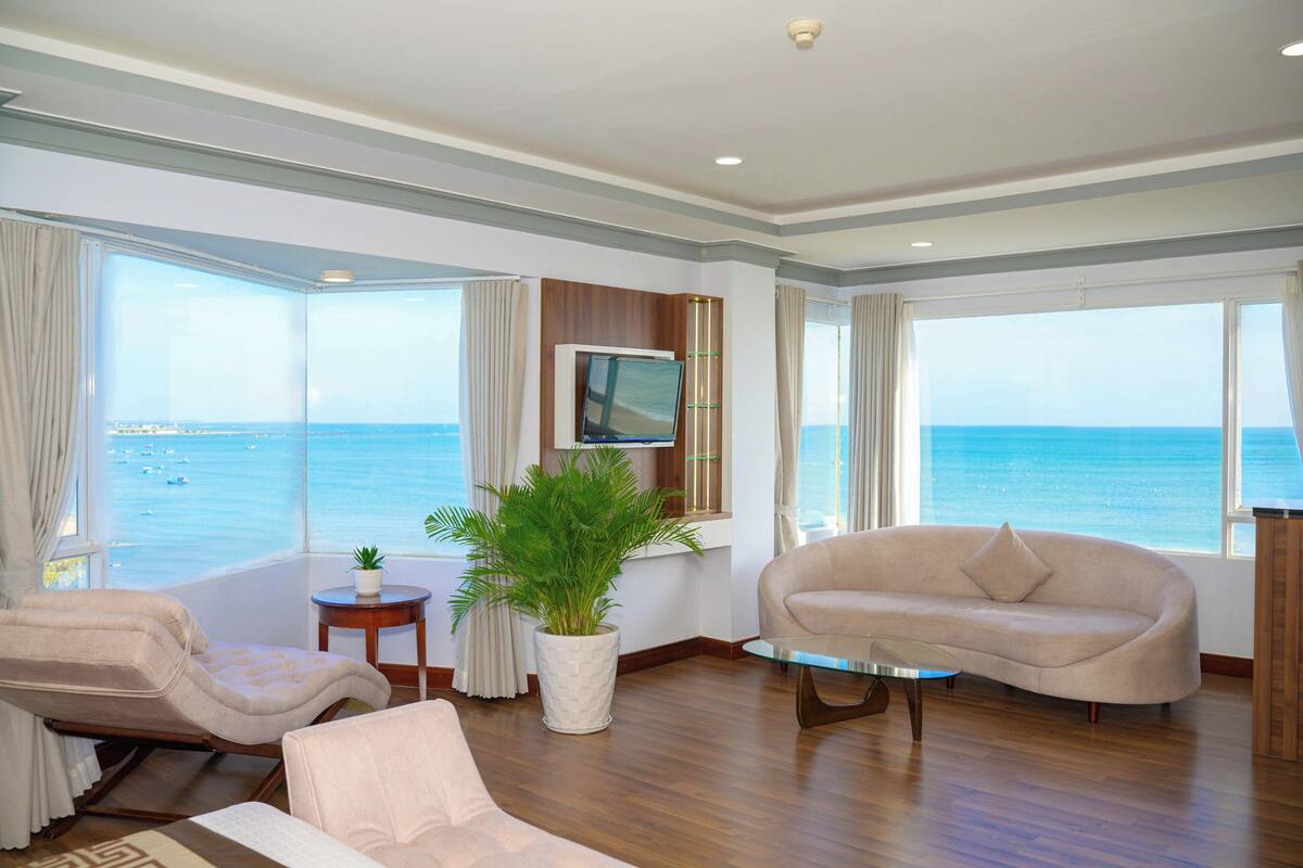 A modern living room with a large window offering a view of the sea, a comfortable sofa, a chair, and a small table with a plant.