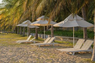 A beach with white lounge chairs and umbrellas, palm trees, and a stone wall in the background.