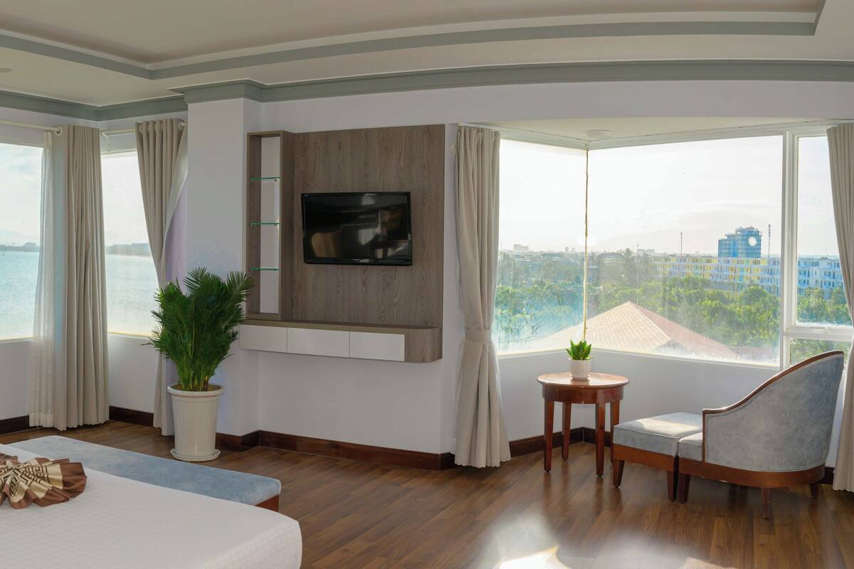 A hotel room with a bed, a TV mounted on the wall, a small table with a plant, and large windows offering a view of the sea and cityscape.