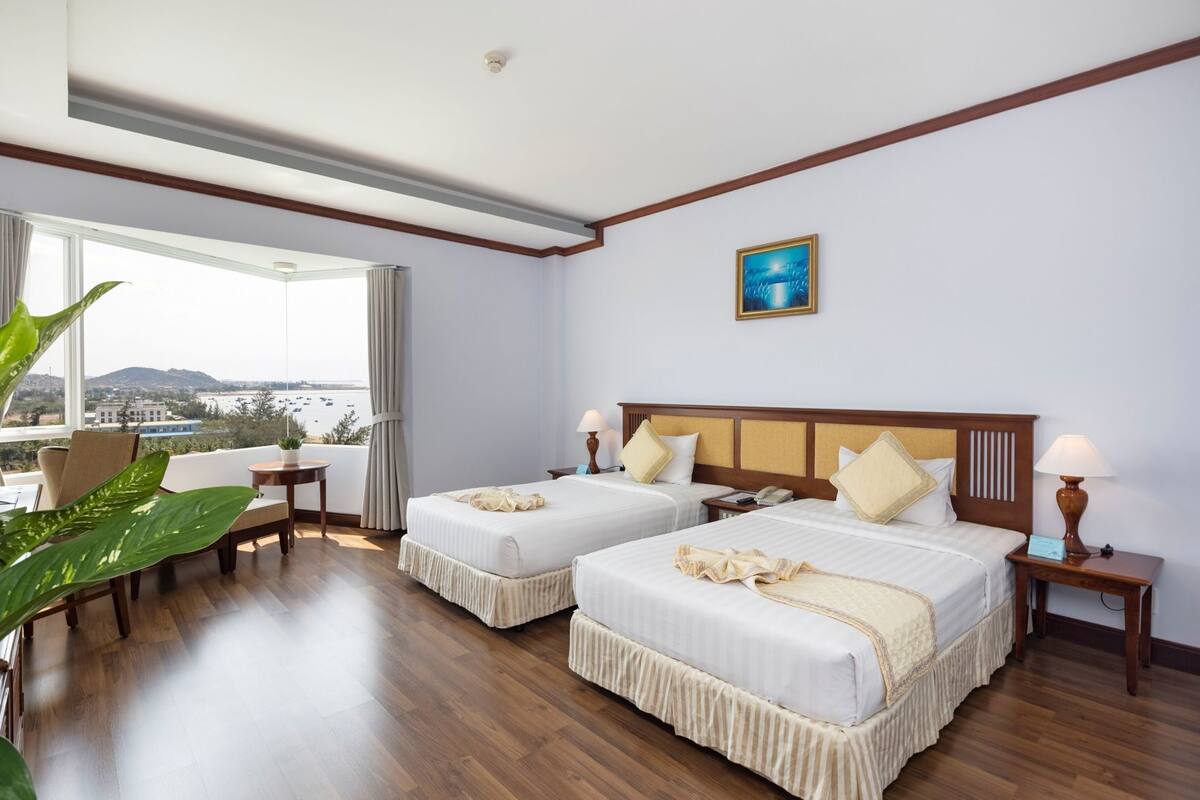 A hotel room with two beds, a large window with a view, a wooden floor, and a painting on the wall.