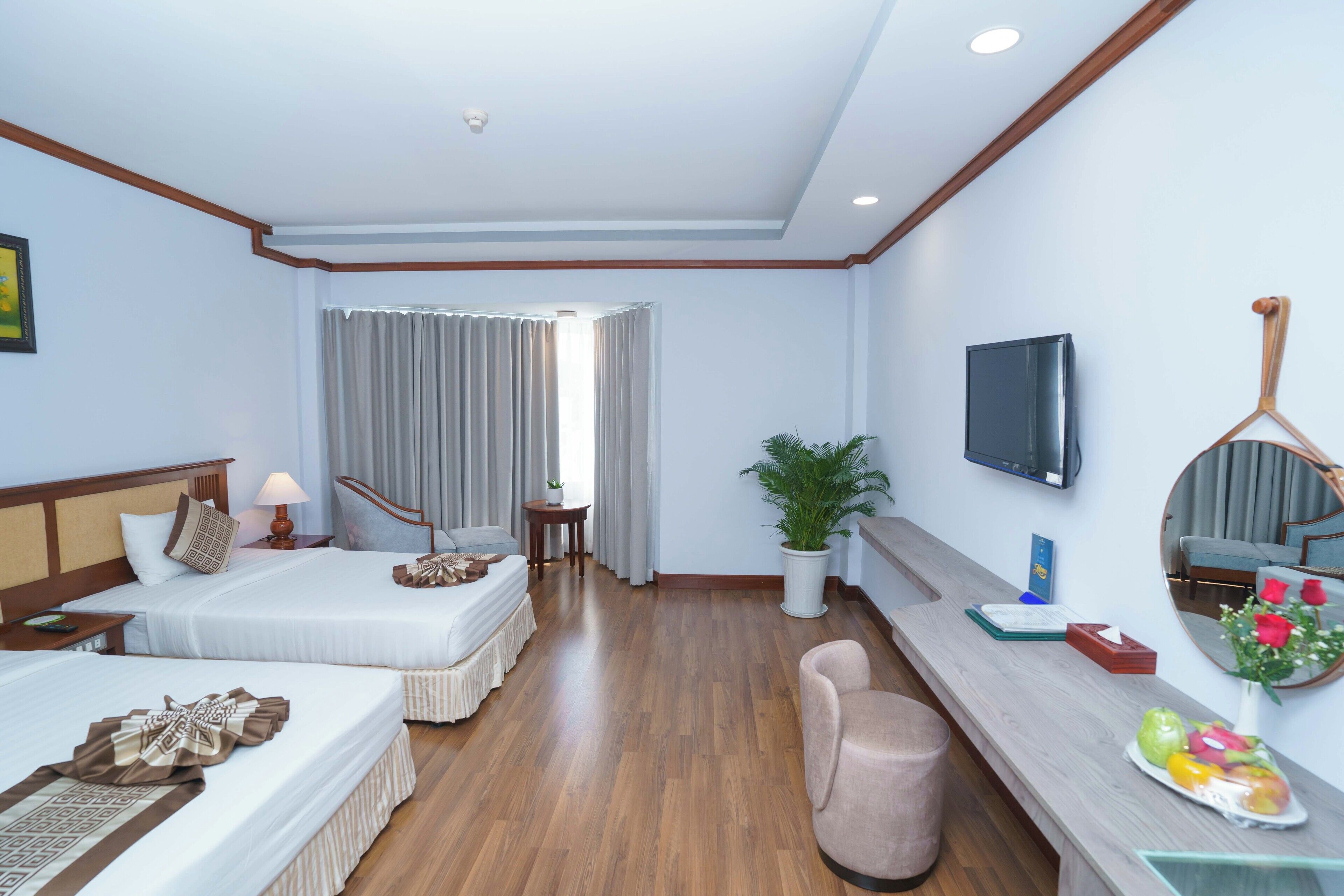 A hotel room with two beds, a TV, a mirror, a plant, and a fruit bowl.