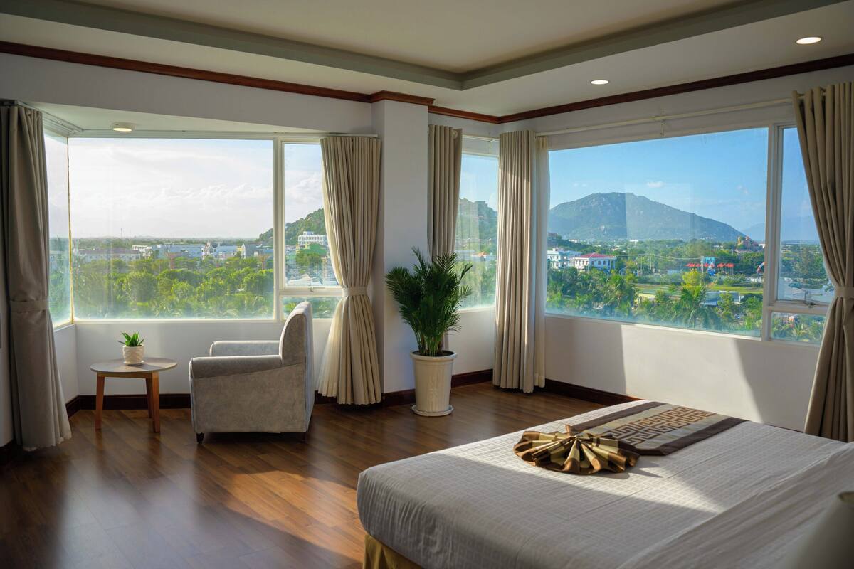 A hotel room with a large bed, a chair, a small table with a plant, and a view of a cityscape and mountains through large windows.