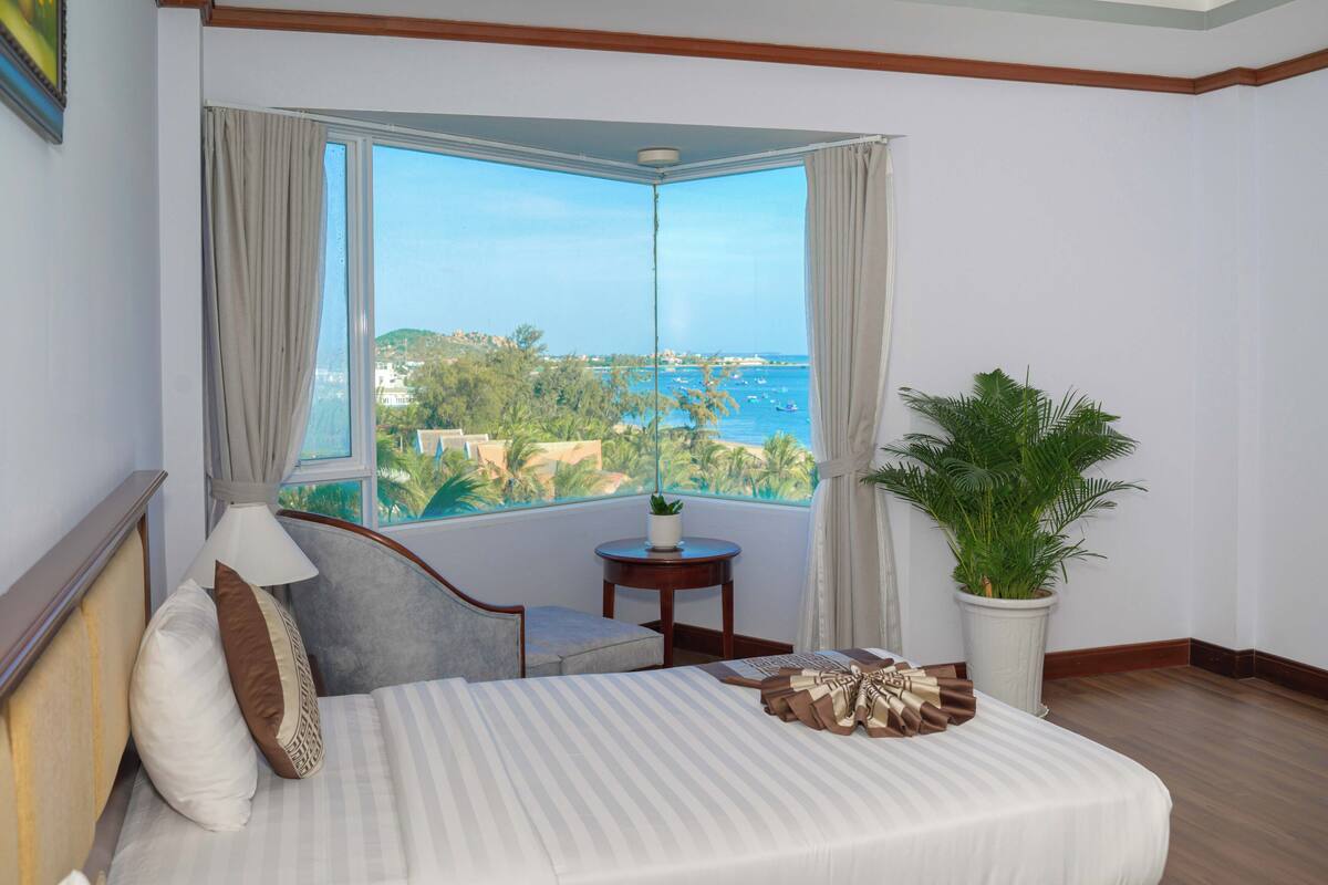 A bedroom with a large window, a bed, a chair, a small table, and a potted plant.