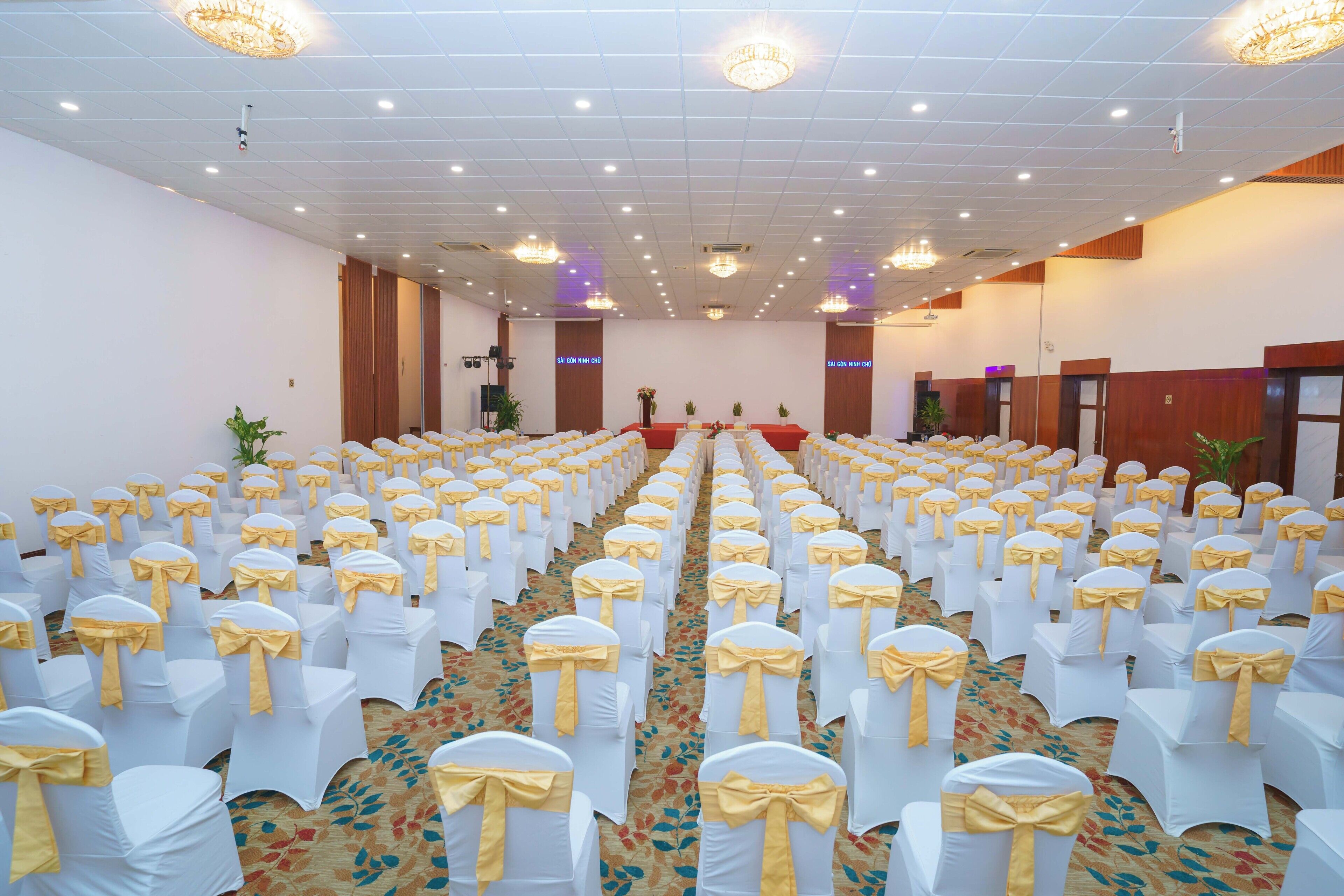 A large hall with rows of white chairs with yellow sashes, a carpeted floor, and chandeliers on the ceiling.