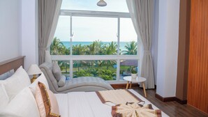 A bedroom with a large window, a bed with pillows, a bedside table, and a view of the ocean and palm trees. - Saigon Ninh Chu Hotel & Resort (Ninh Chu)