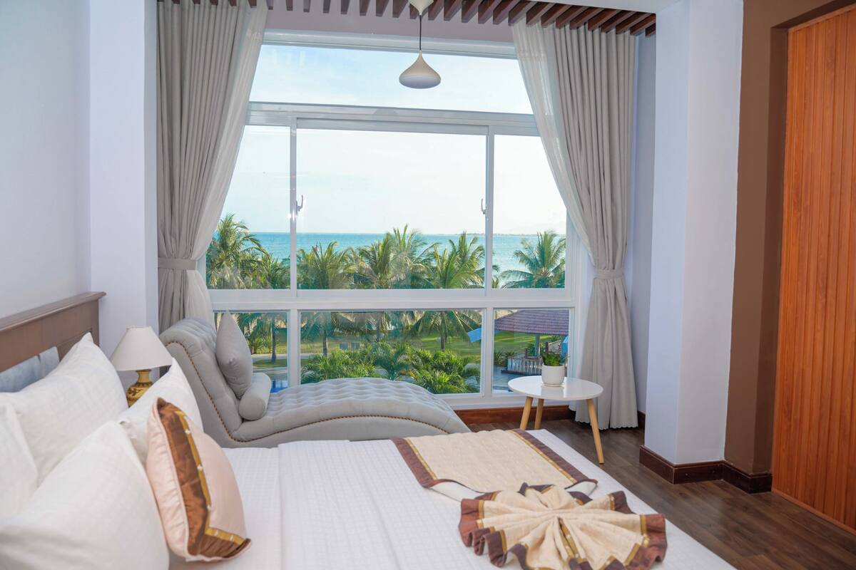 A bedroom with a large window, a bed with pillows, a bedside table, and a view of the ocean and palm trees.