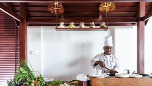 A chef preparing food in a kitchen with wooden beams, a shelf with jars, and a variety of fresh ingredients and dishes on the counter. - Kingdom Angkor Hotel (Siem Reap)