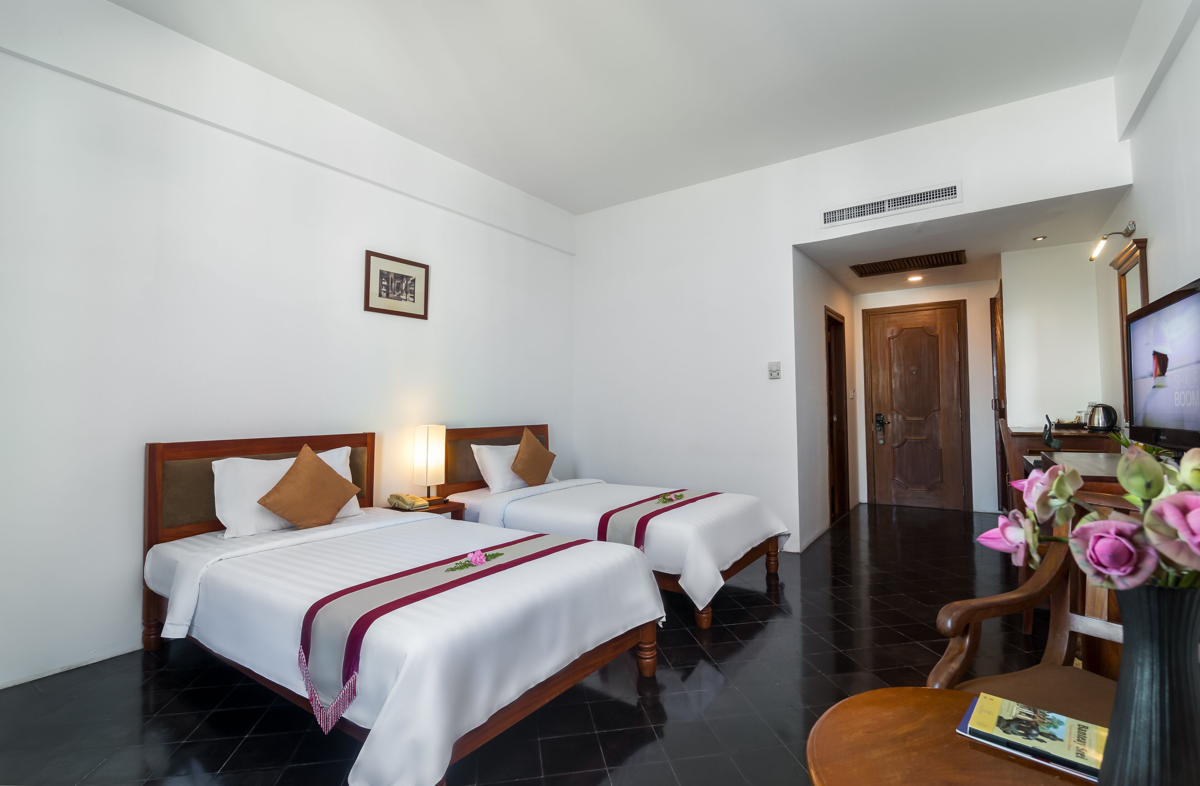 A hotel room with two beds, a wooden headboard, a nightstand with a lamp, a wooden chair, a table with a book, and a vase with flowers.