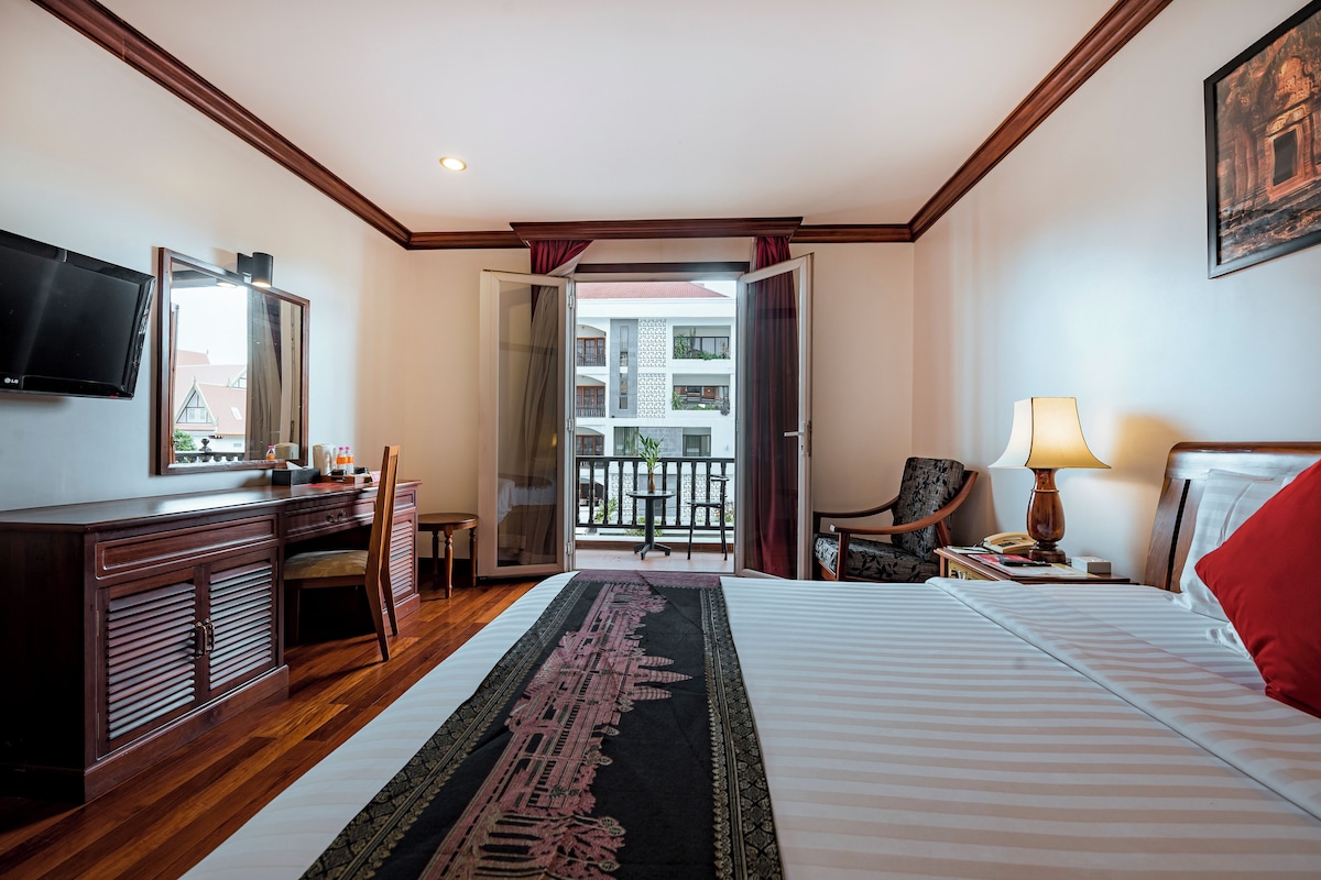 A hotel room with a bed, a desk, a chair, a TV, a balcony, and a lamp.