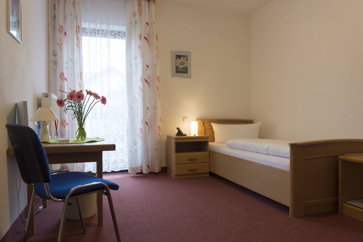 A hotel room with a bed, a desk with a chair, a nightstand, a lamp, and a vase with flowers.