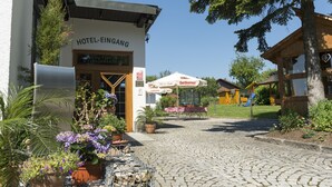 A hotel entrance with a cobblestone path, outdoor seating, and a playground in the background. - Hotel Dreisonnenberg (Neuschoenau)