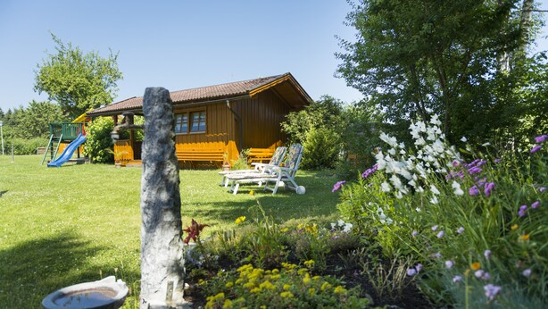 A wooden cabin with a slide in the yard, surrounded by a garden with various flowers and a birdbath. - Hotel Dreisonnenberg (Neuschoenau)