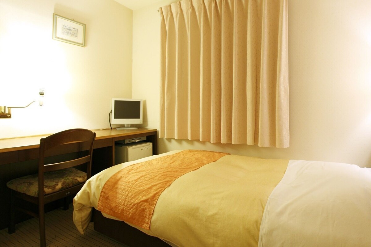 A hotel room with a bed, a desk with a computer, a chair, and a window with curtains.