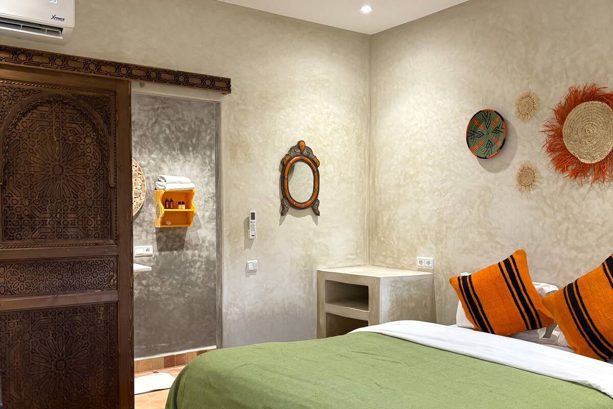 Un dormitorio con una colcha verde, una puerta de madera con intrincados grabados, una mesita de noche pequeña y un espejo fijado a la pared.