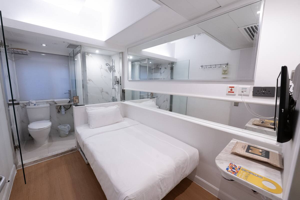 A modern hotel room with a white bed, a glass shower enclosure, a white toilet, and a large mirror.