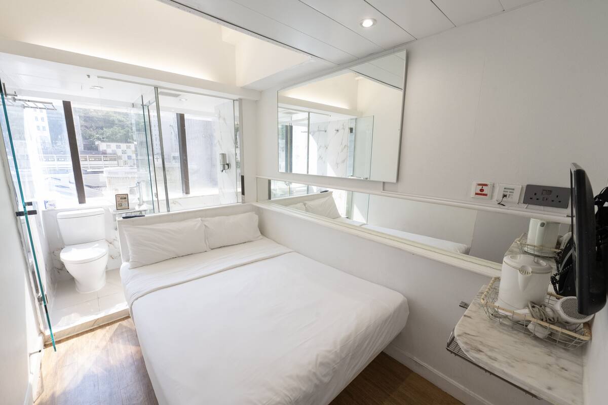 A modern hotel room with a large bed, a bathroom with a toilet and shower, a large window, and a marble countertop with a coffee maker.