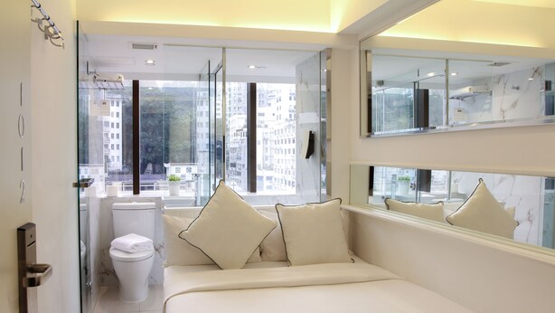 Habitació d'hotel moderna amb una dutxa de vidre, un vàter blanc, un llit gran amb almoades beix i un mirall gran que reflecteix l'habitació.
