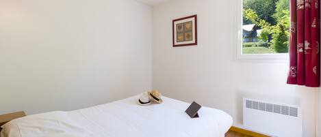 Una cama bien hecha con ropa blanca, una mesita de noche con un ordenador portátil, un cuadro en la pared y una ventana con vistas a la vegetación.