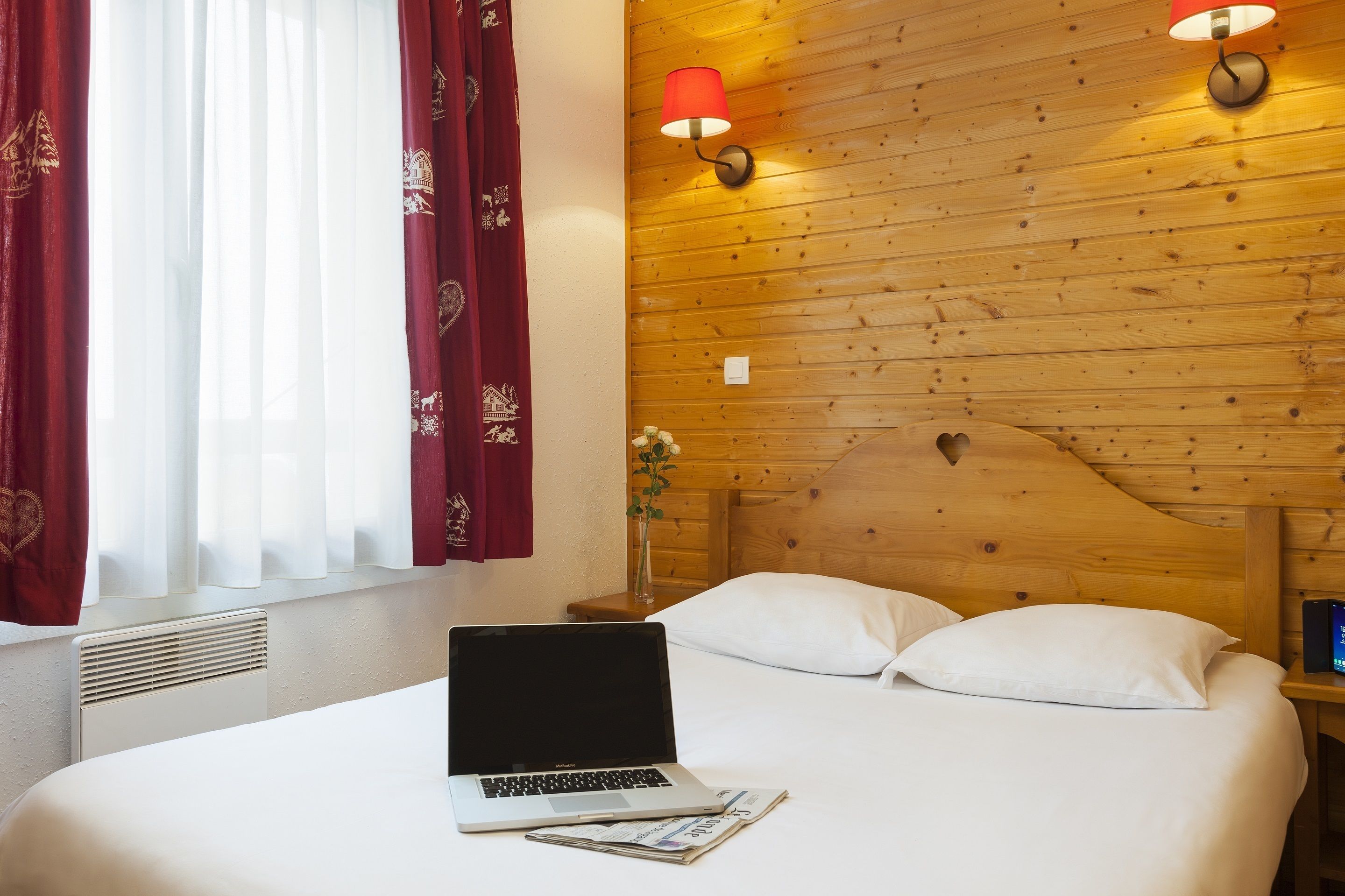 A hotel room with a wooden headboard, a bed with white linens, a laptop on the bed, and a nightstand with a lamp.