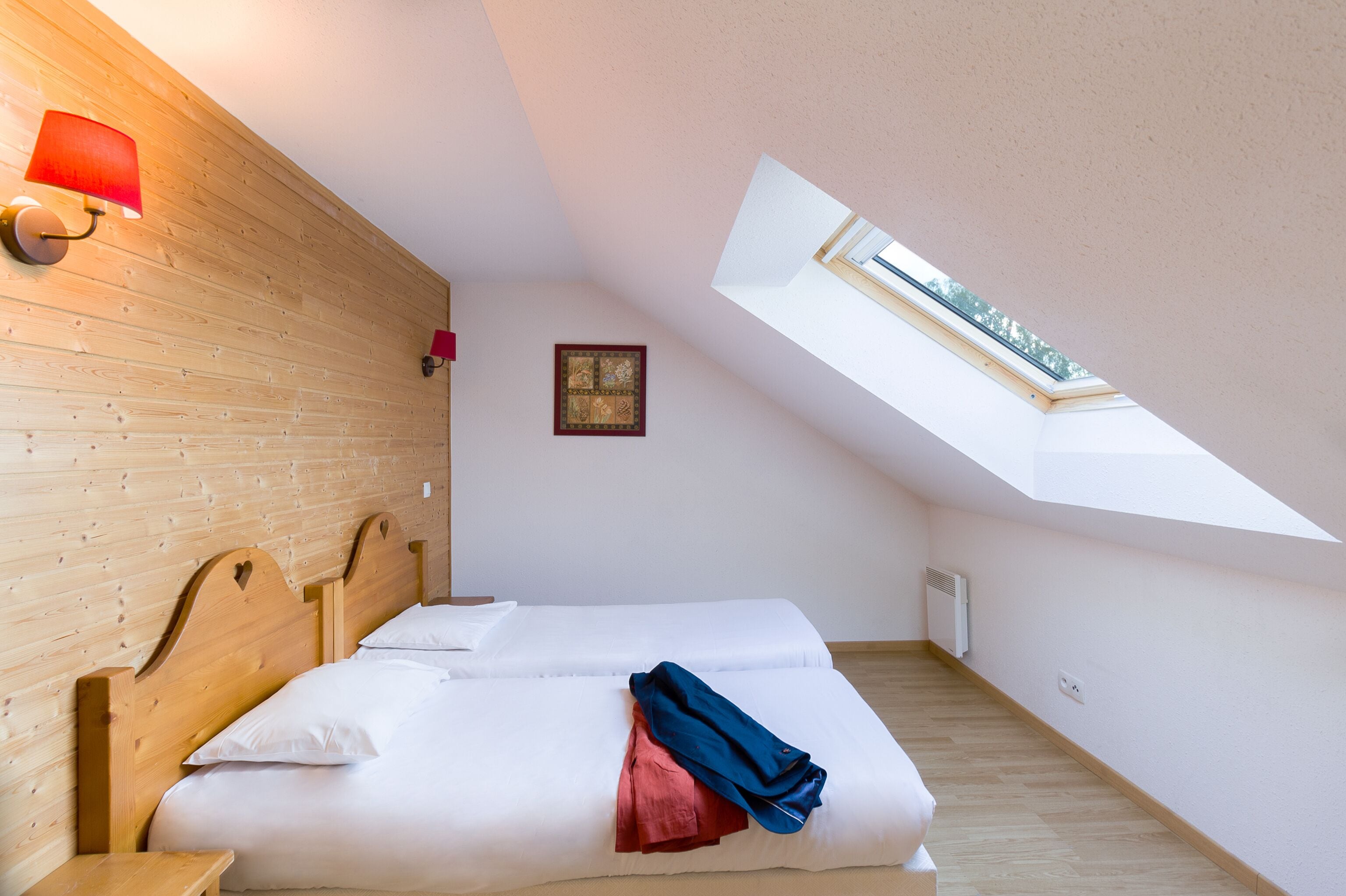 Una habitación con techo inclinado, tragaluz, cabecera de madera, dos camas, una lámpara roja y un cuadro en la pared.