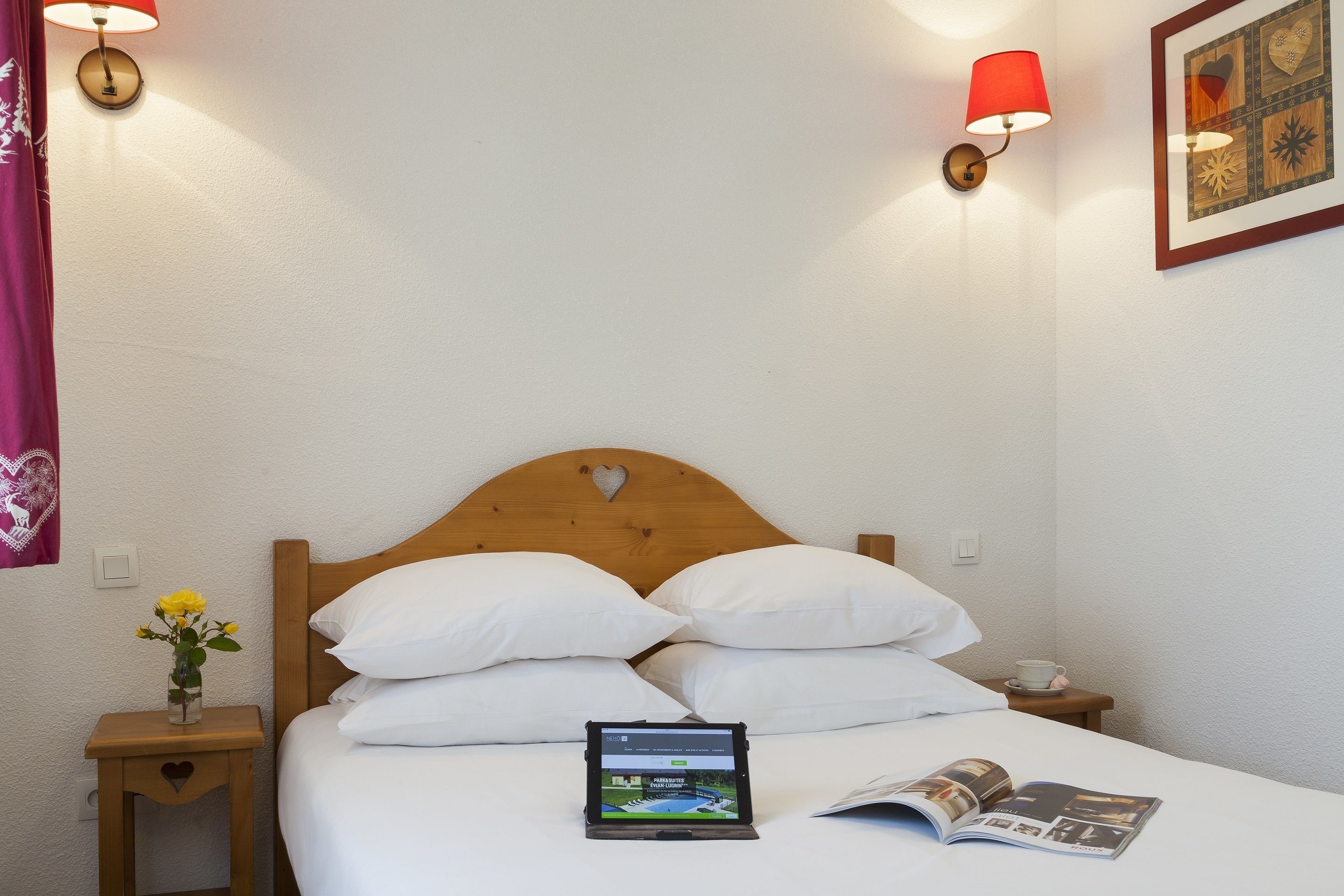 A neatly made bed with white linens, a tablet on the bed, and a magazine open on the bed.