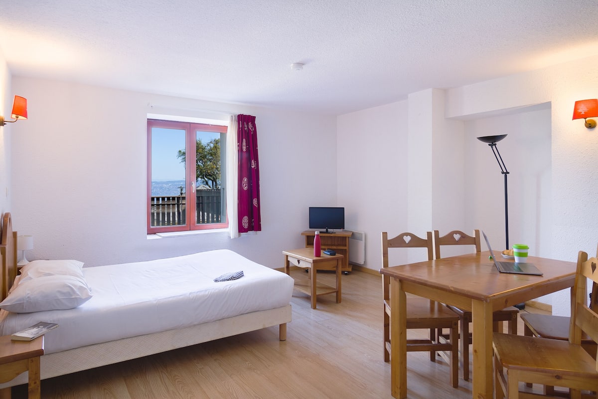 A hotel room with a bed, desk, chair, table, TV, and a view of the outdoors.
