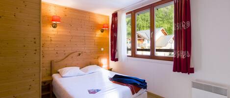 Una habitación con paneles de madera, una cama, una ventana con cortinas y una lámpara fijada a la pared.