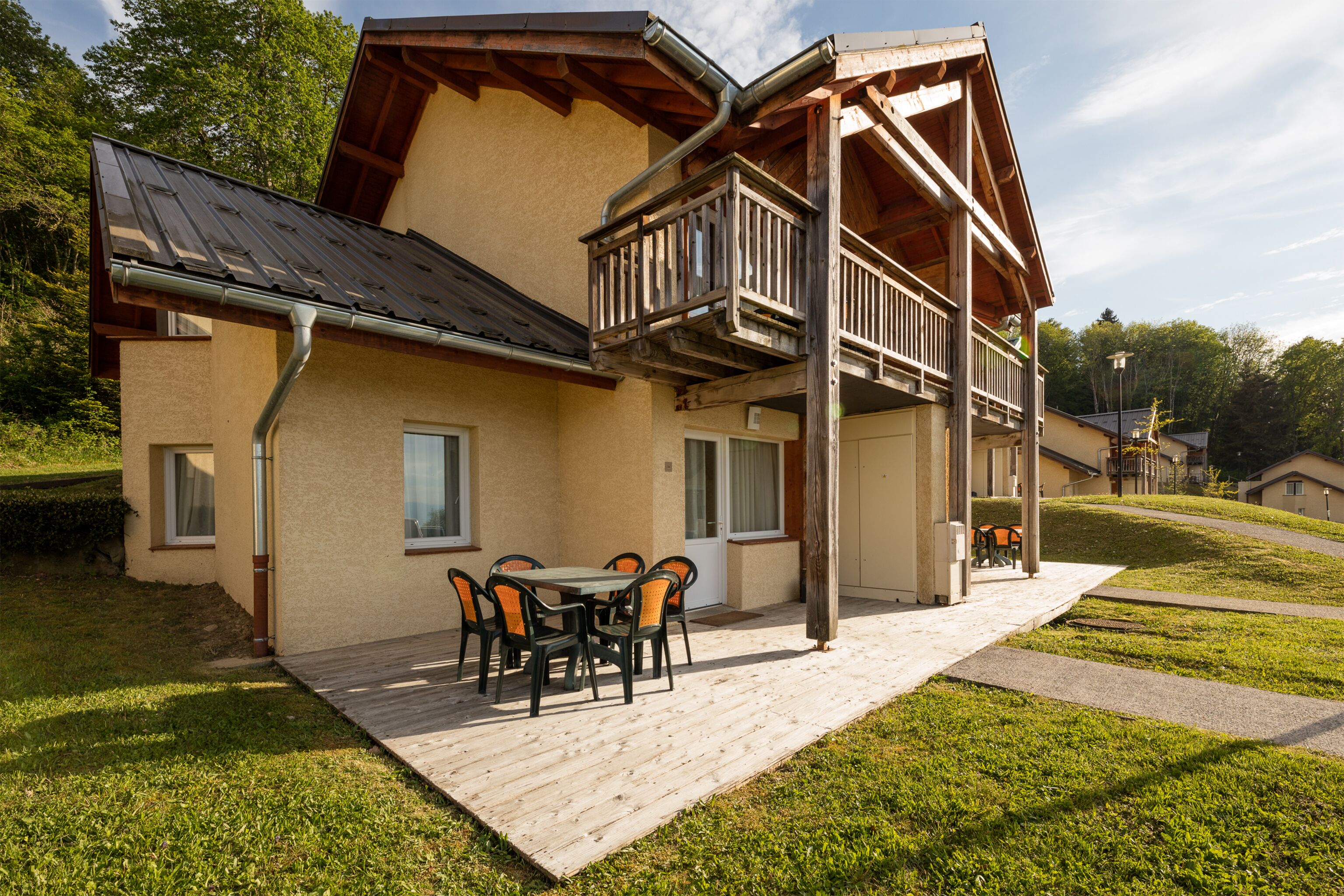 Een gebouw met twee verdiepingen, een houten balkon, een terras met tafel en stoelen, en een gazon.