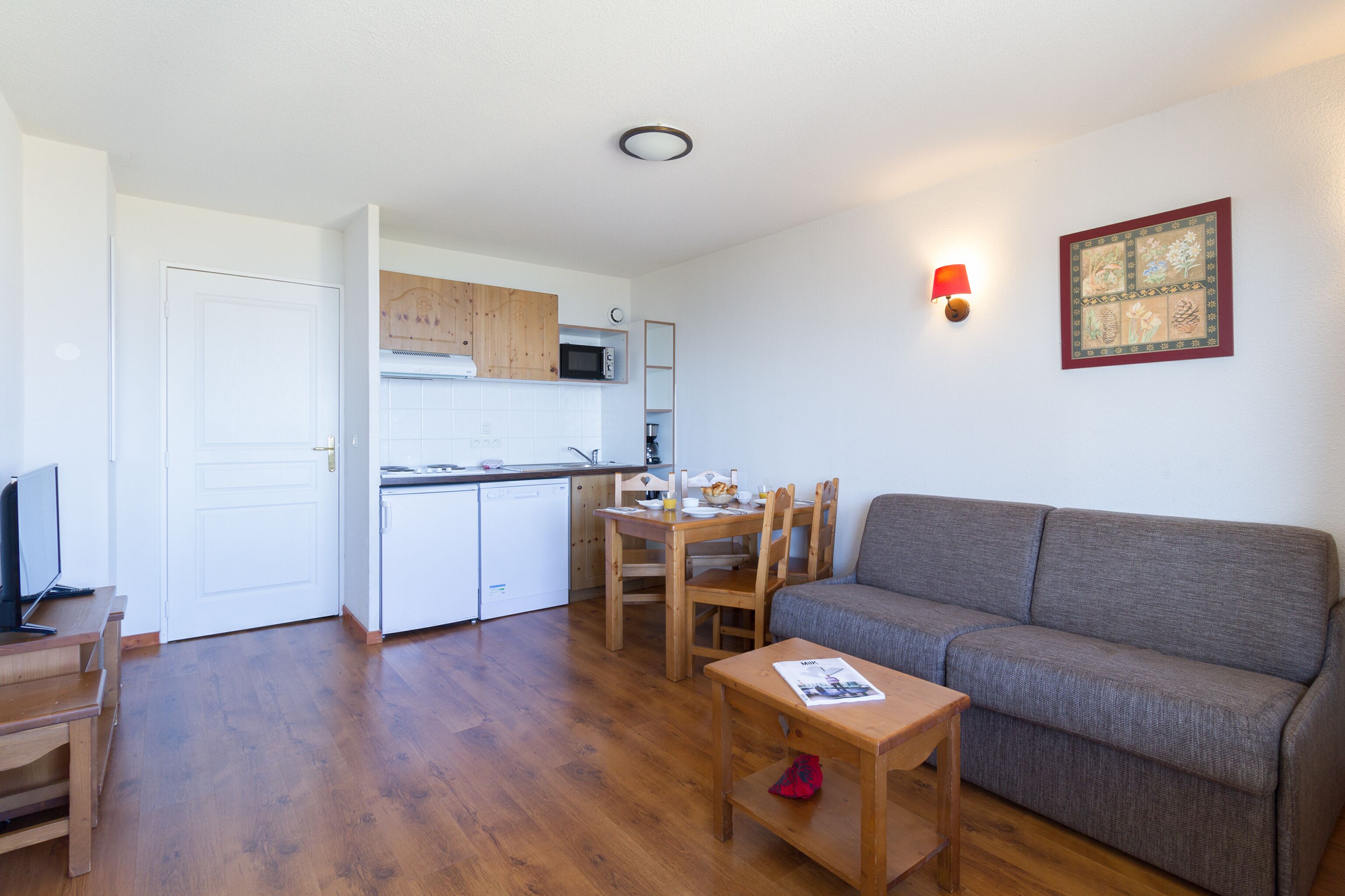 A compact living space with a kitchenette, a gray sofa, wooden furniture, and a wall-mounted light fixture.