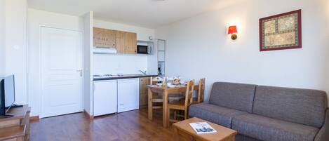 Un espacio habitable compacto con kitchenette, un sofá gris, muebles de madera y una lámpara de pared.