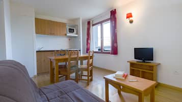 Quarto compacto com cama, mesa de jantar com cadeiras, televisão em suporte e cozinha equipada com armários e micro-ondas.