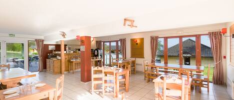A spacious dining area with multiple wooden tables and chairs, a kitchen area, and large windows providing natural light.
