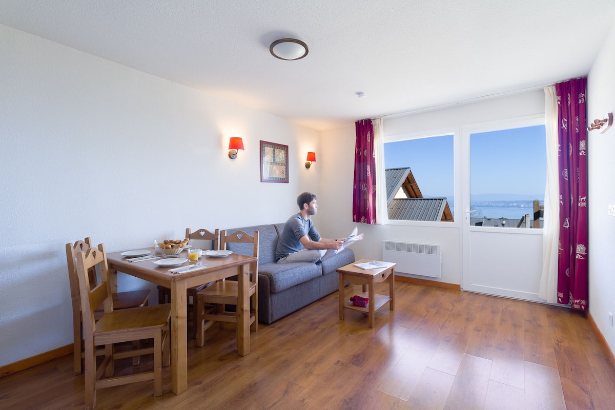 A room with a dining table, chairs, a sofa, a small table, and a window with a view of a building and the sky.