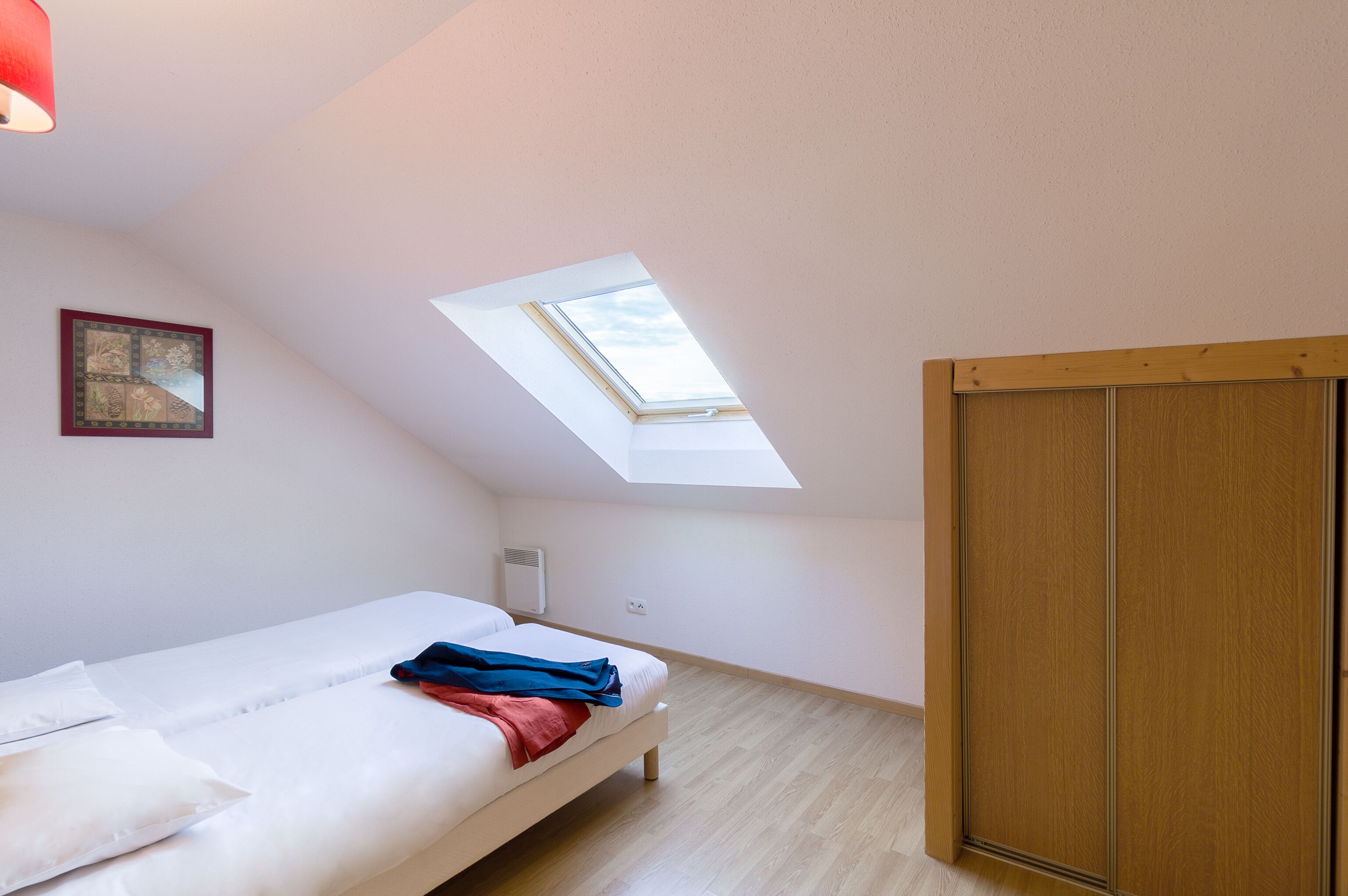 Een slaapkamer met dakraam, een bed, een houten kledingkast en een ingelijst schilderij aan de muur.