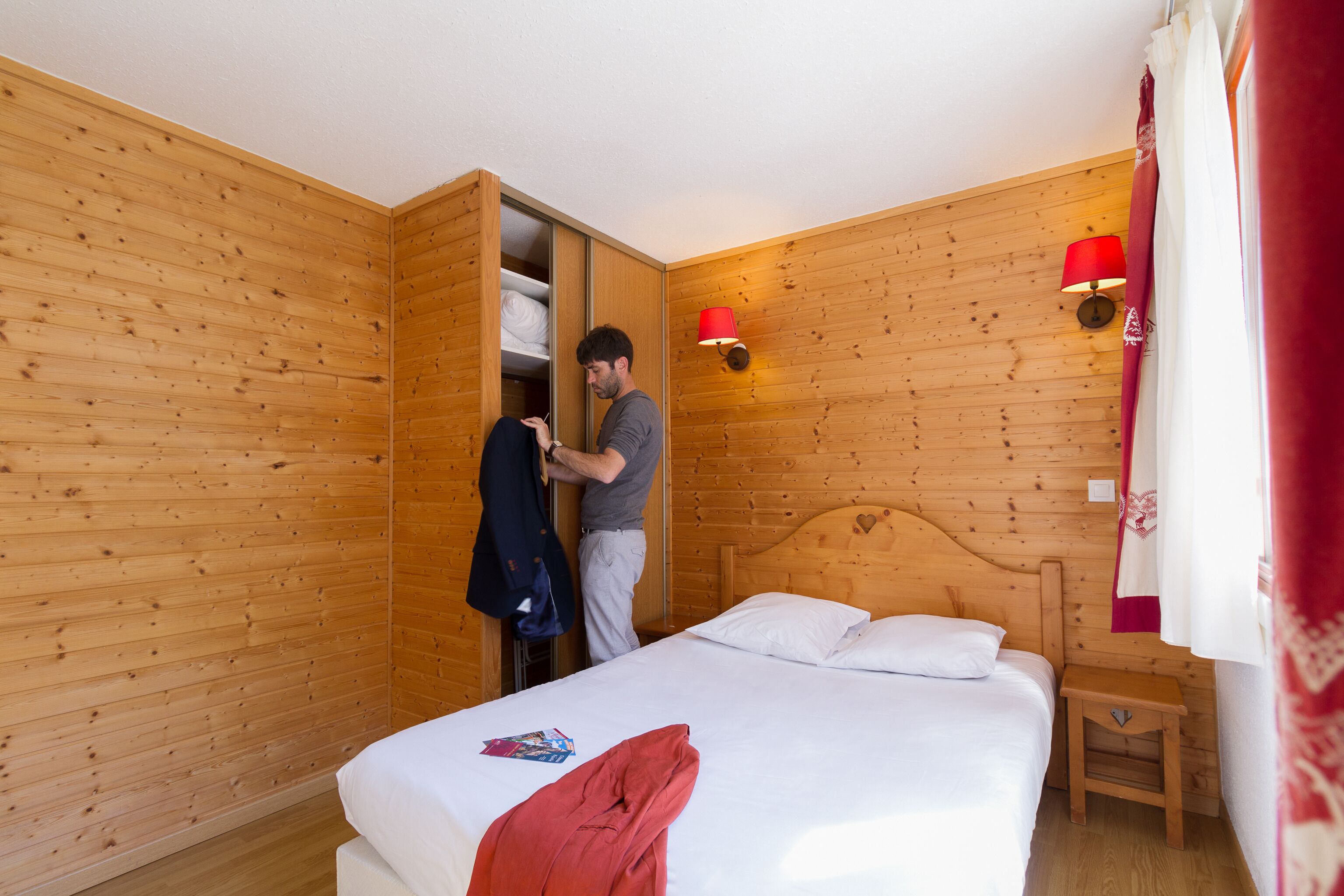A wooden-paneled room with a bed, a nightstand, a red lamp, and a wardrobe.