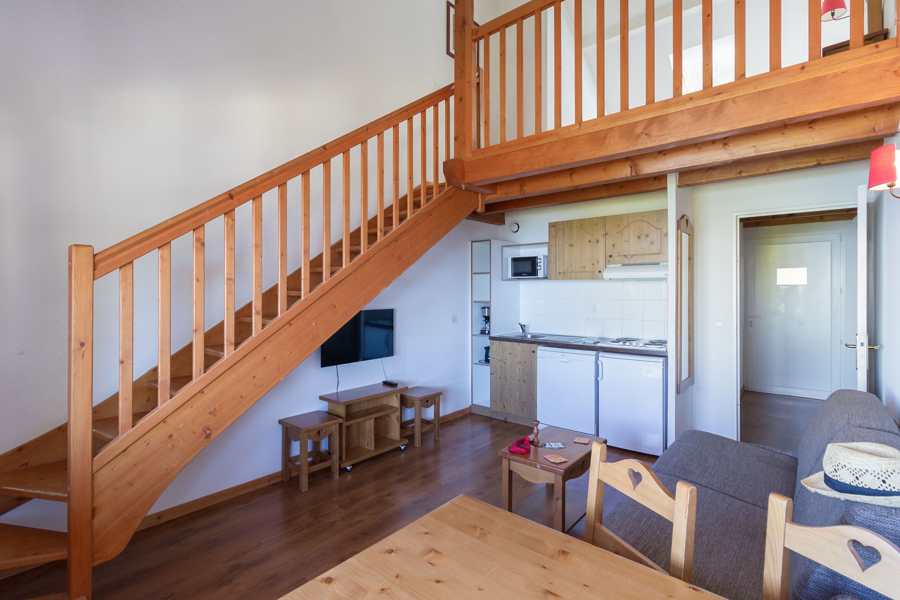 Un escalier en bois menant à un niveau supérieur, un coin cuisine avec des meubles blancs, une table à manger avec des chaises et un canapé sur lequel est posé un chapeau.