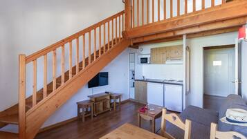 Una escalera de madera que conduce a un nivel superior, una zona de cocina con armarios blancos, una mesa de comedor con sillas y un sofá con un sombrero.
