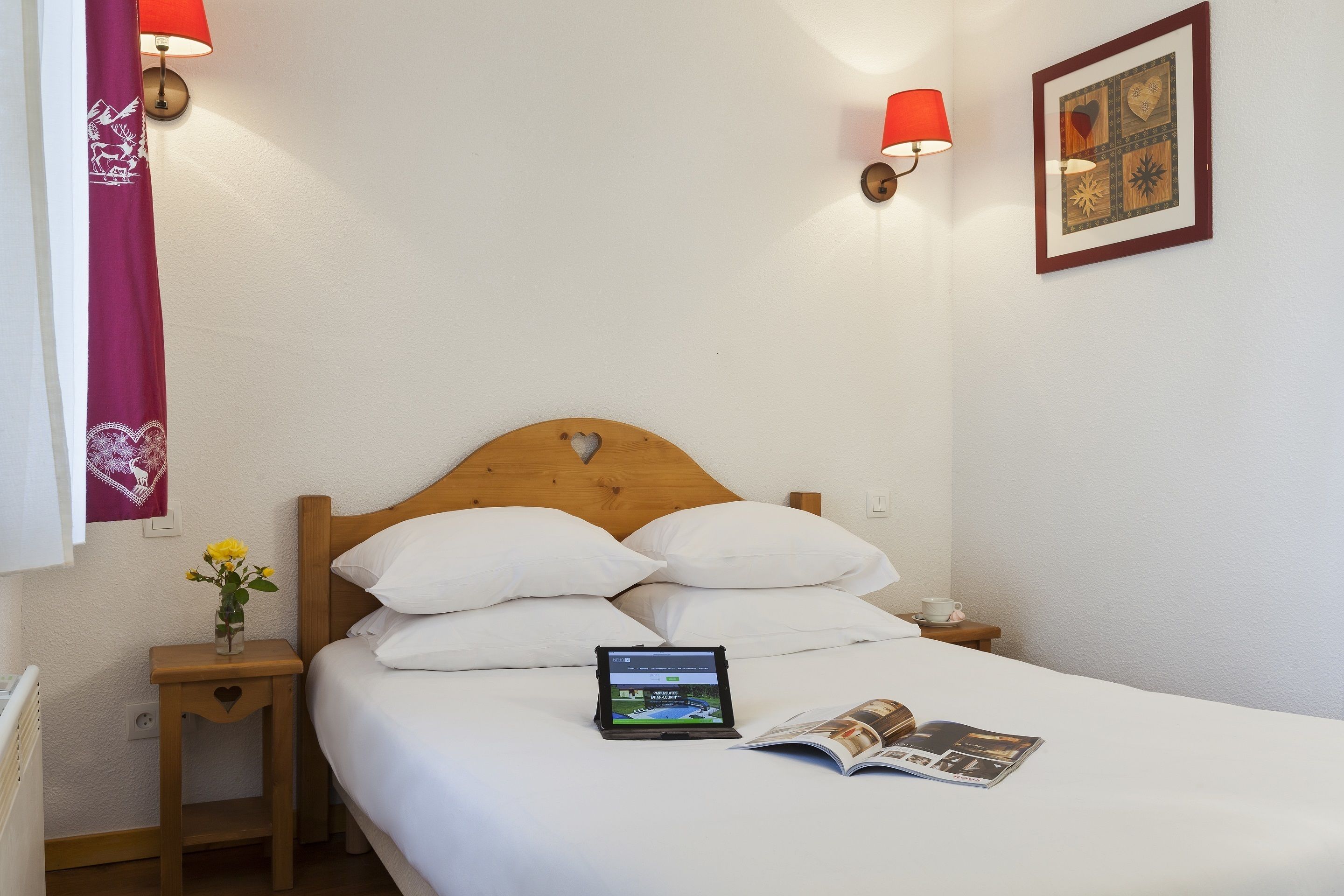 A neatly made bed with white linens, a tablet on the bed, and a magazine beside it. There is a small bedside table with a vase of flowers and a lamp.