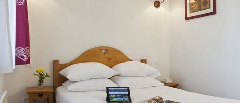 A neatly made bed with white linens, a tablet on the bed, and a magazine beside it. There is a small bedside table with a vase of flowers and a lamp.